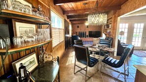 Een houten interieur met een eethoek, bestaande uit een glazen tafel en stoelen, een bar met een plank vol glazen, en een televisie op de achtergrond.