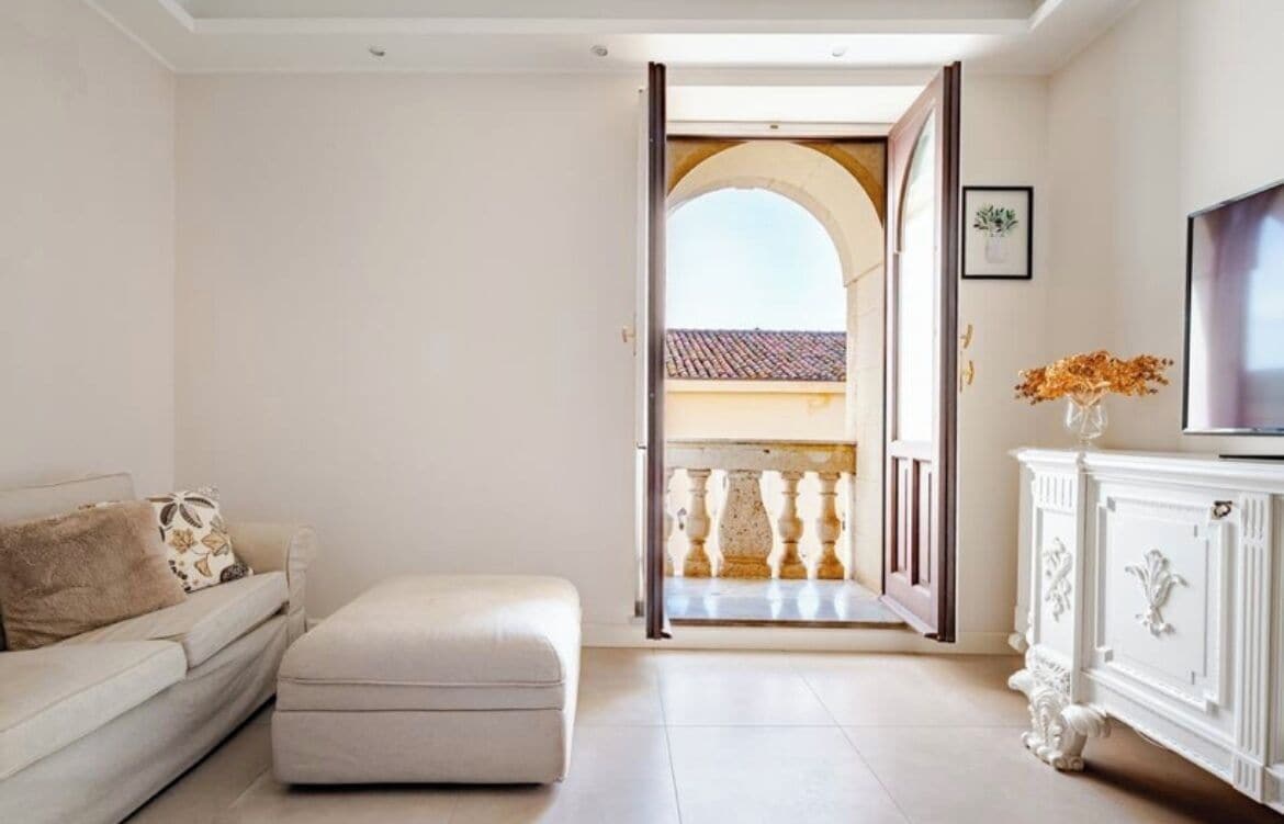 A room with a white sofa, a matching ottoman, a white cabinet, and a TV. An open door leads to an outdoor balcony with a railing.