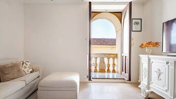 A room with a white sofa, a matching ottoman, a white cabinet, and a TV. An open door leads to an outdoor balcony with a railing.