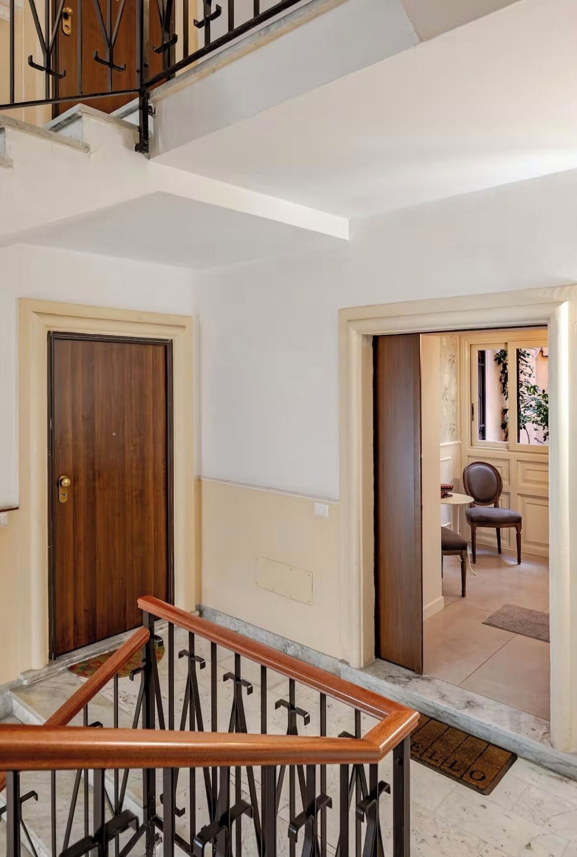 Una escalera con barandilla de madera y balaustres de metal, que conduce a una habitación con puerta de madera y una pequeña mesa con una silla.