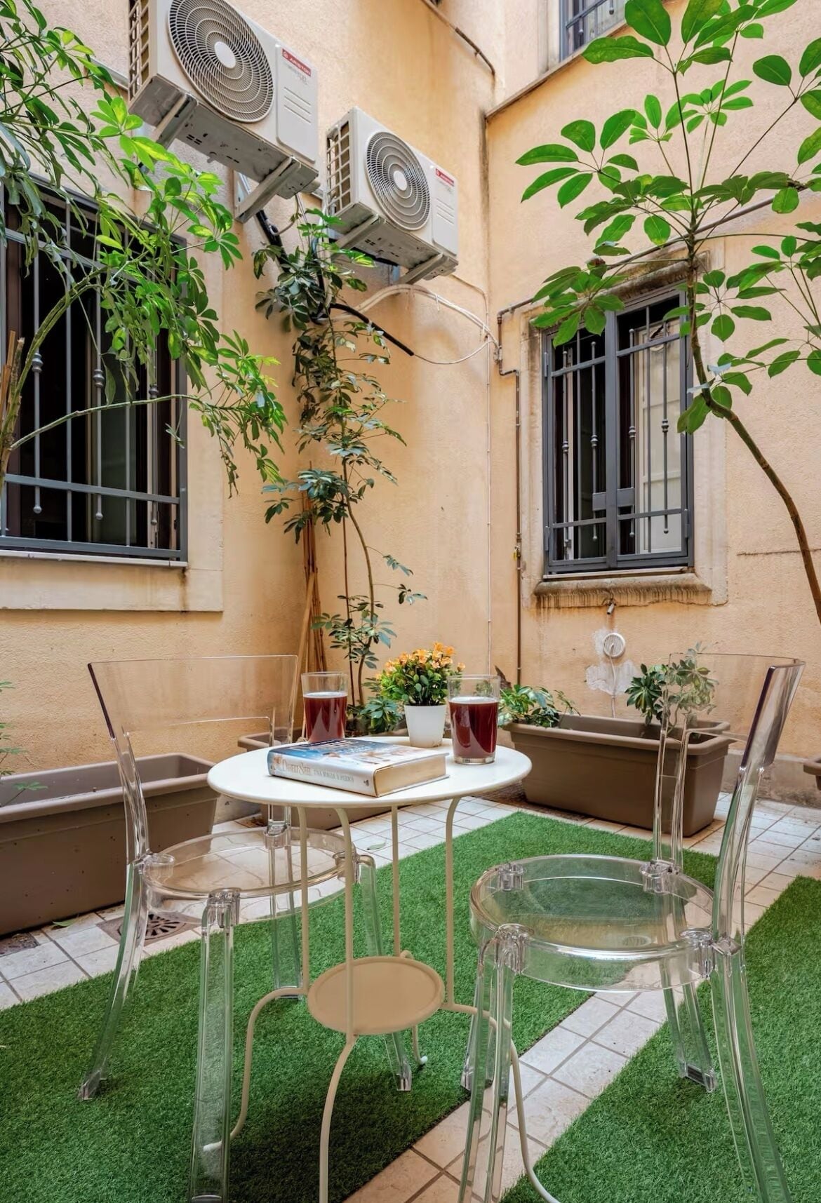 Une petite terrasse avec une table ronde, des chaises transparentes et du gazon synthétique. Des unités de climatisation sont fixées au mur, et des plantes en pot sont disposées tout autour.