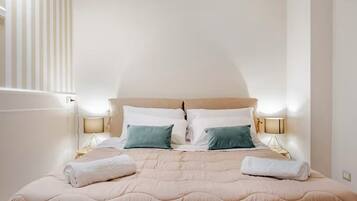 Una cama bien hecha con una colcha color beige y almohadas, dos lámparas de noche y una ventana con persianas verticales.
