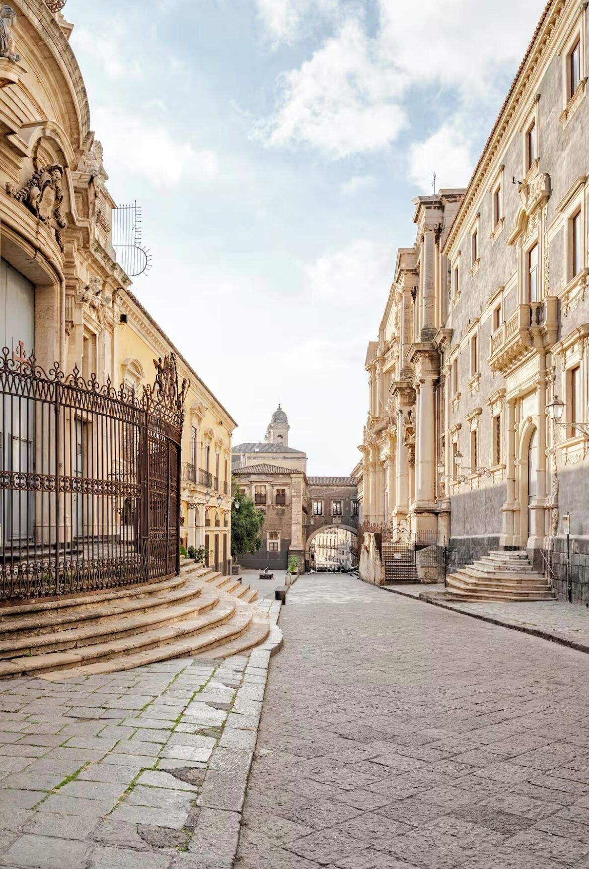 Una calle adoquinada flanqueada por edificios históricos con fachadas ornamentadas y un arco central que conduce a una estructura con cúpula.