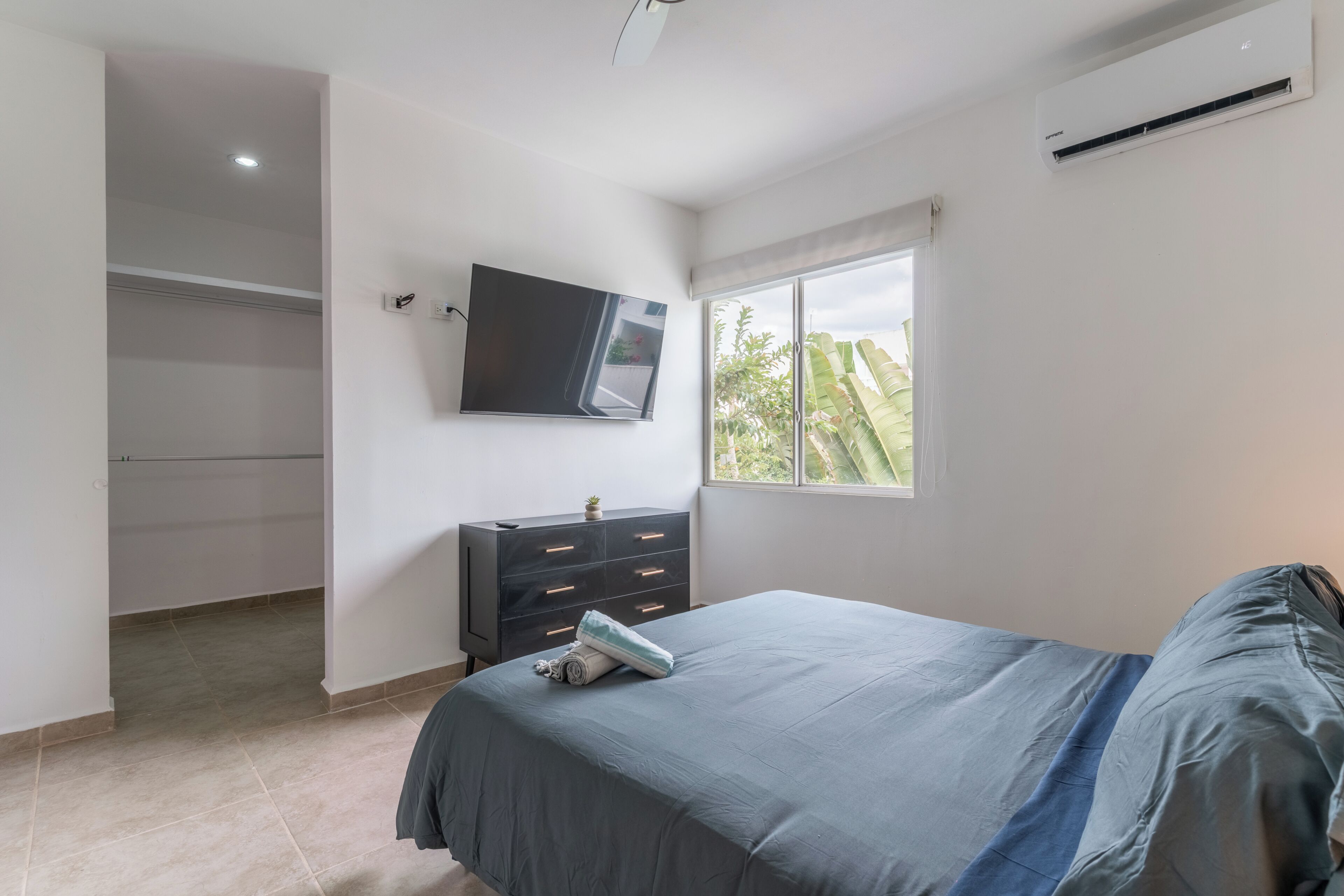Een slaapkamer met een bed, een dressoir, een aan de muur bevestigde televisie en een raam met uitzicht op groen.