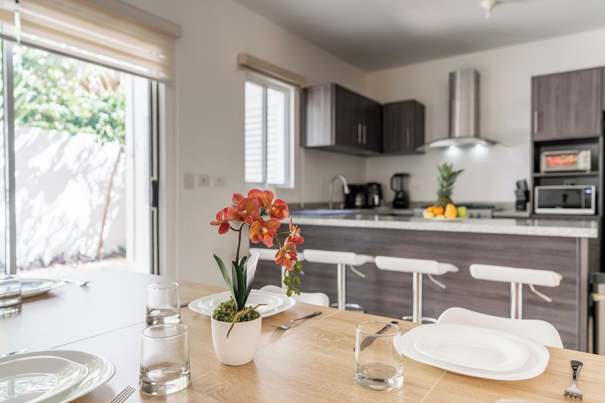 A dining area with a table set for two, a potted orchid, and a kitchen in the background.