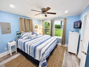 A bedroom with a bed, a ceiling fan, a window with curtains, a bedside table, and a TV on the wall. - Clown Fish Cottage Rear, King Bed, 1 Block to Beach (Mexico Beach)
