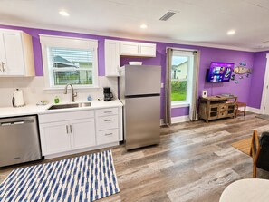 A compact kitchen with white cabinets, a stainless steel refrigerator, and a wooden dining table. - Clown Fish Cottage Rear, King Bed, 1 Block to Beach (Mexico Beach)
