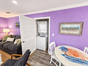 Ein Zimmer mit einer violetten Wand, einer weißen Tür, einem grauen Sofa, einem runden Tisch mit bunten Tischdecken und einem weißen Stuhl.