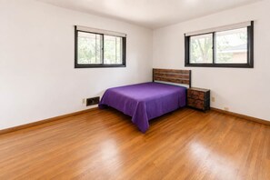 Ein Schlafzimmer mit Holzboden, einem Bett mit violetter Bettdecke, zwei Fenstern und einem Nachttisch.