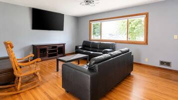 A living room with a sofa, a wooden rocking chair, a coffee table, and a flat-screen TV mounted on the wall.