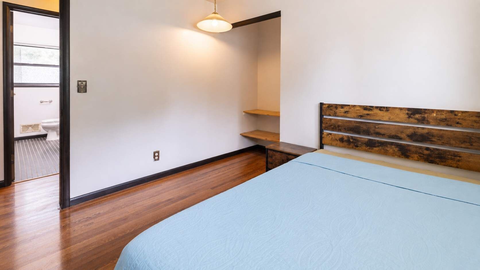 A bedroom with a bed, a wooden headboard, a hanging light fixture, and a wooden floor.