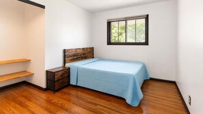 Ein Schlafzimmer mit Bett, Nachttisch, Holzboden, weißen Wänden und einem Fenster mit Blick auf Bäume.