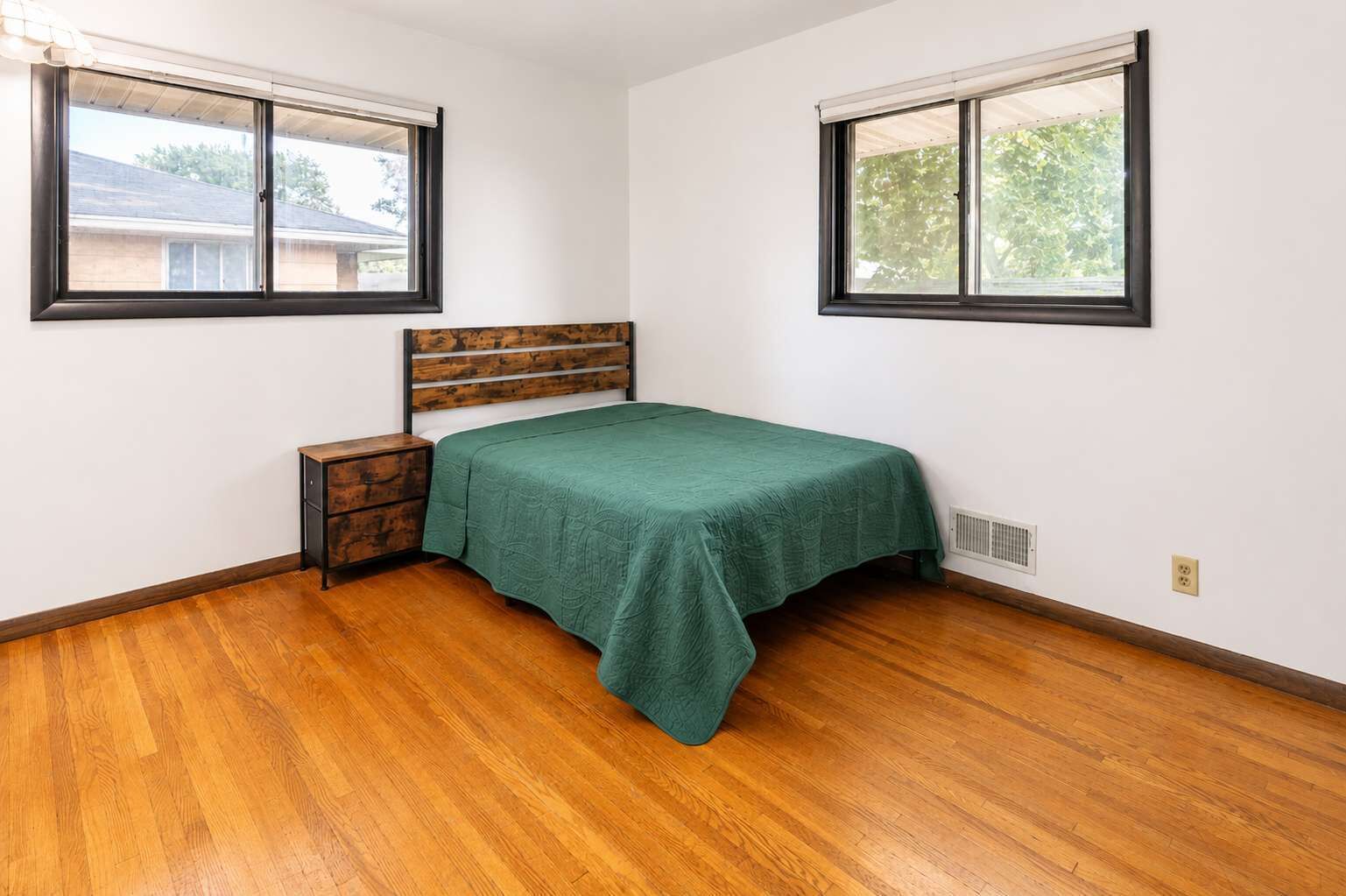 Un dormitorio con cama, mesita de noche de madera, dos ventanas y suelo de parqué.