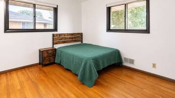 A bedroom with a bed, wooden nightstand, two windows, and hardwood flooring.
