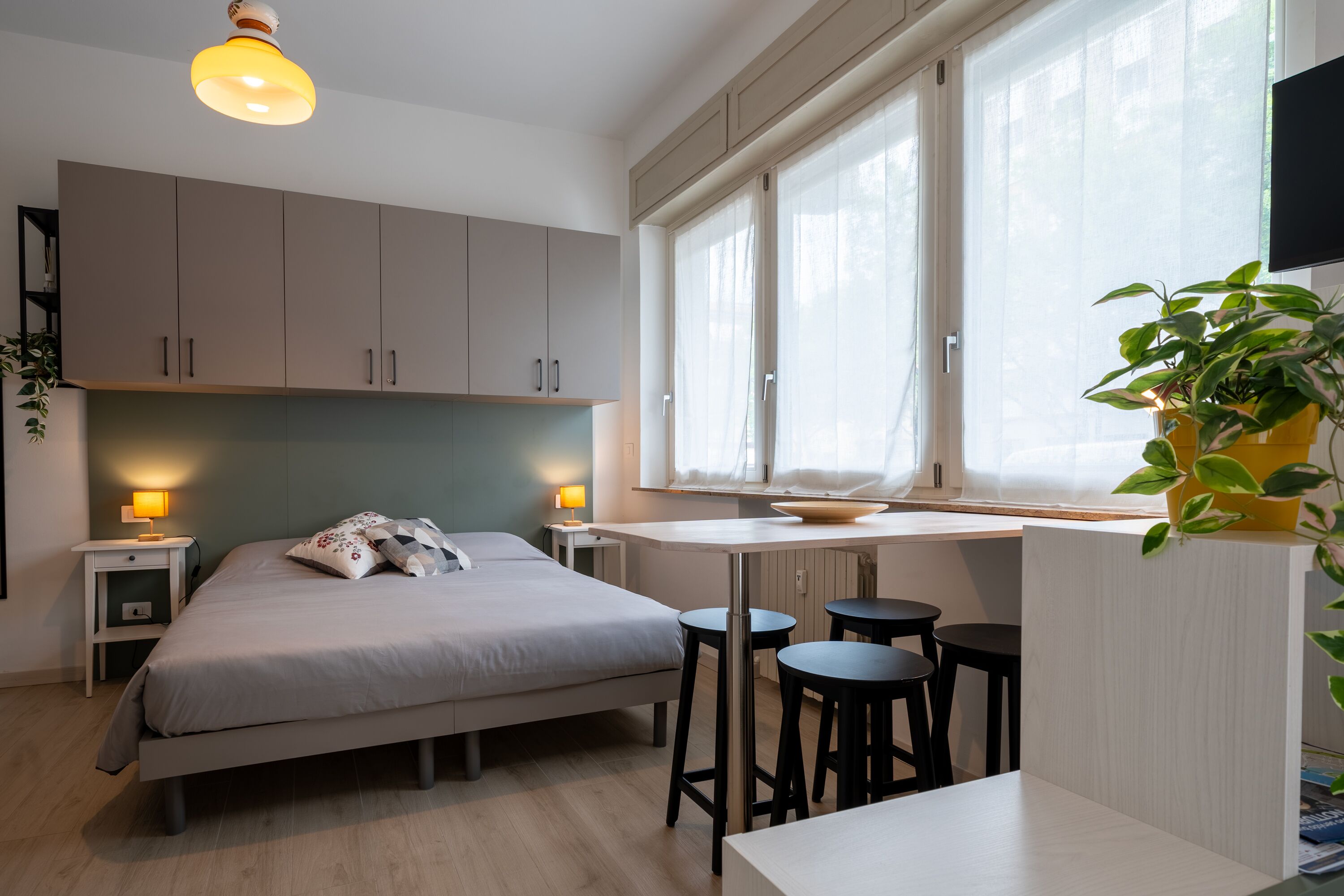 A modern bedroom with a bed, bedside lamps, a desk, and a window with sheer curtains.