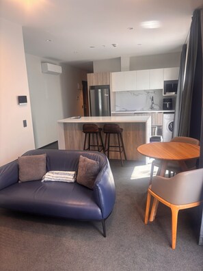 A modern living room with a dark blue sofa, a wooden coffee table, and a small round table with chairs. A kitchen area is visible in the background with a refrigerator, microwave, and washing machine. - Spacious Accessible CBD Apartment, metres from new stadium, free carpark onsite (Christchurch)