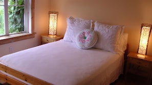 A bed with white linens, two bedside tables with lamps, and a window with greenery outside.