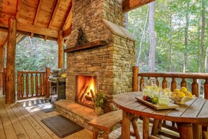 A wooden deck with a stone fireplace, a table with drinks and fruit, and a view of the forest. - 2 King Ensuites on first level | Private with easy access | Outdoor Fireplace | Hot Tub | Game Room (Sevierville)