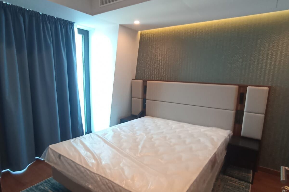 A hotel room with a large bed, a headboard with built-in lighting, a window with curtains, and a small bedside table.