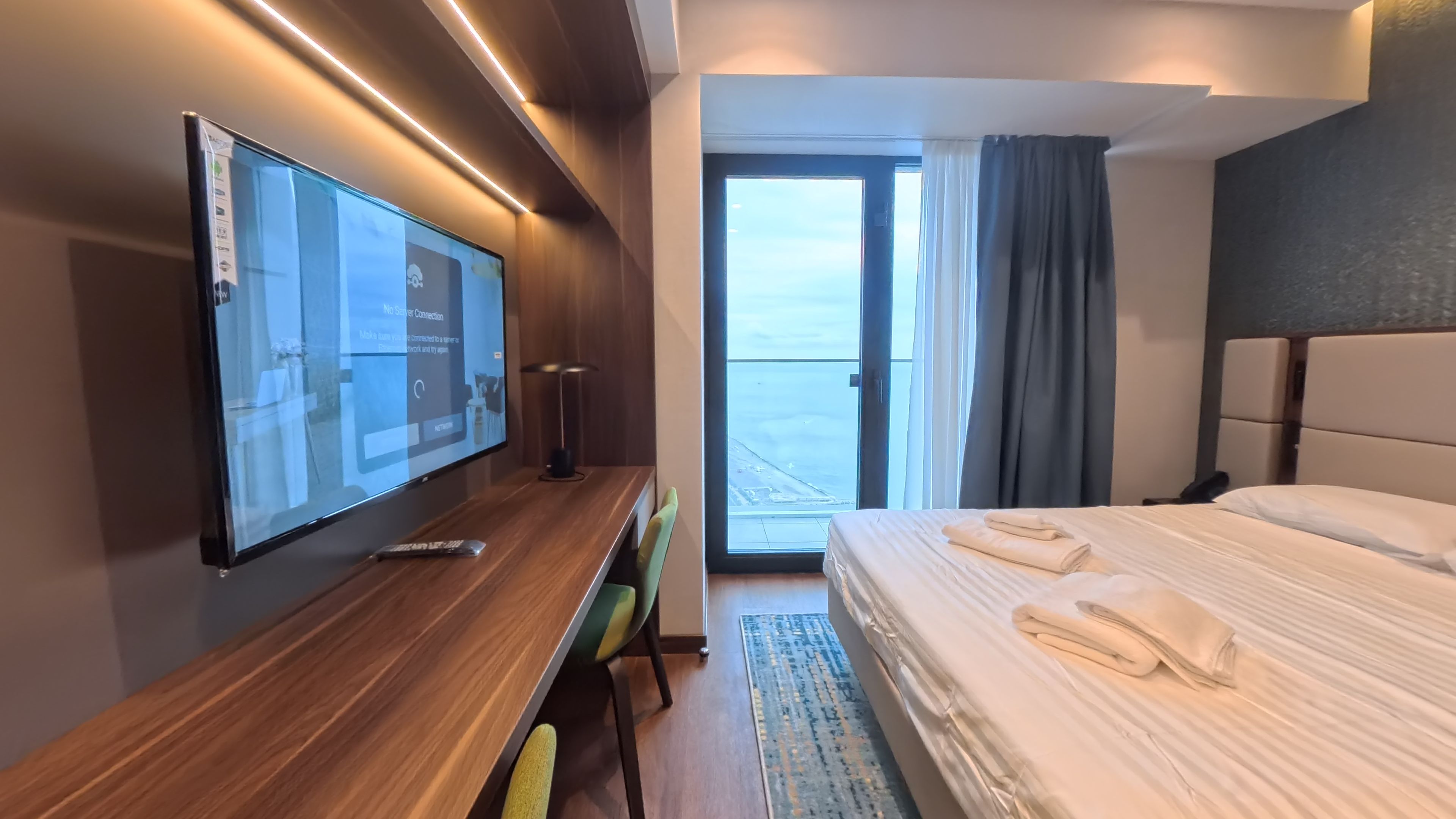 A hotel room with a large flat-screen TV, a wooden desk, a comfortable bed with neatly folded towels, and a balcony with a view.