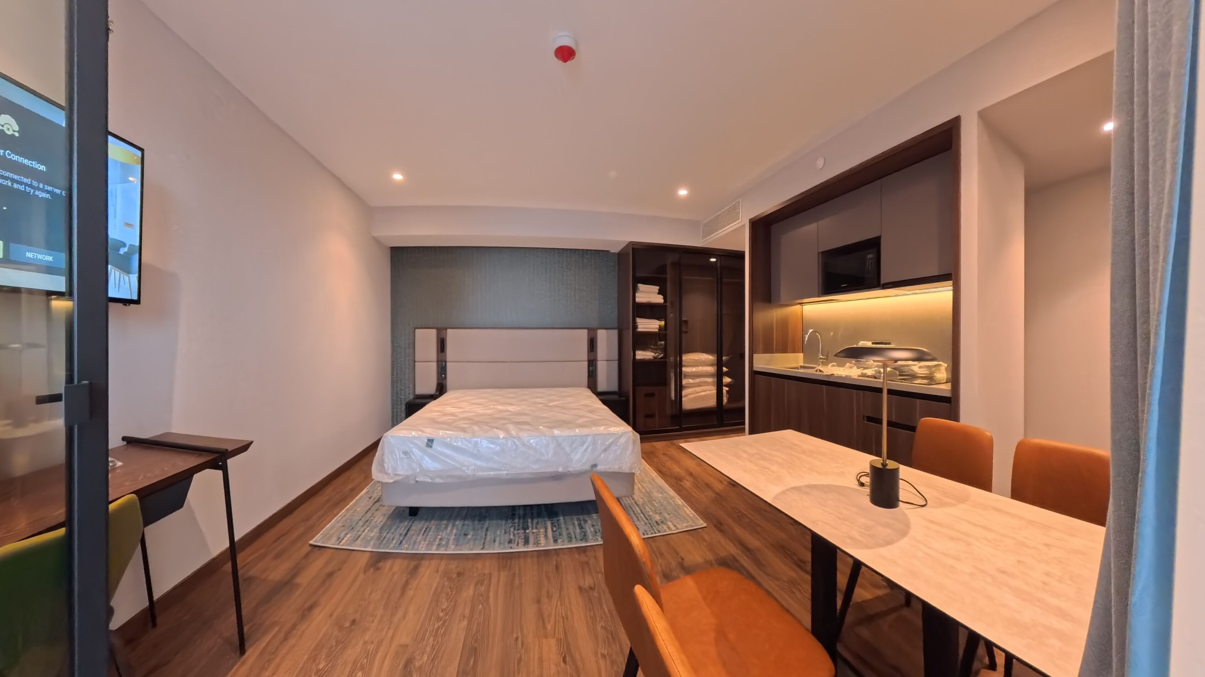 A modern hotel room with a bed, desk, chair, and a kitchenette area.