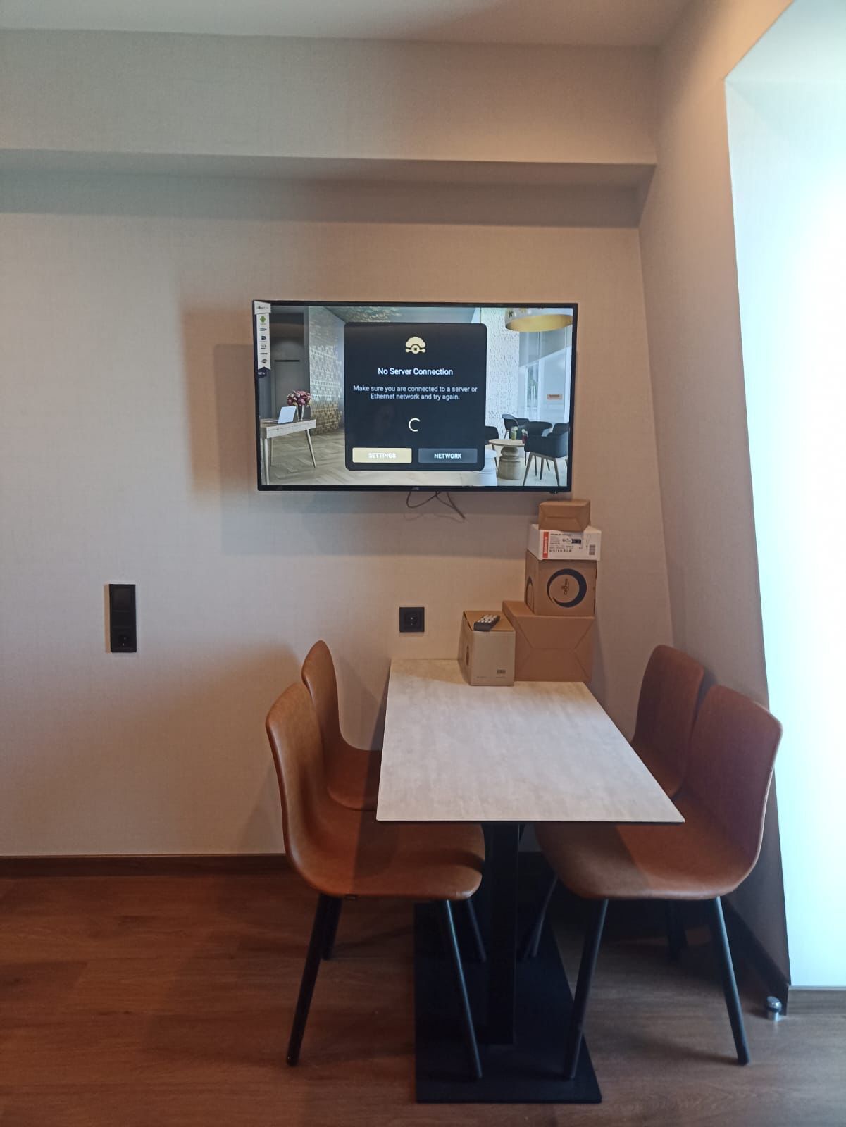 A modern living room with a flat-screen TV mounted on the wall, a dining table with two chairs, and a small speaker on the table.