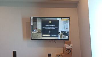 A modern living room with a flat-screen TV mounted on the wall, a dining table with two chairs, and a small speaker on the table.