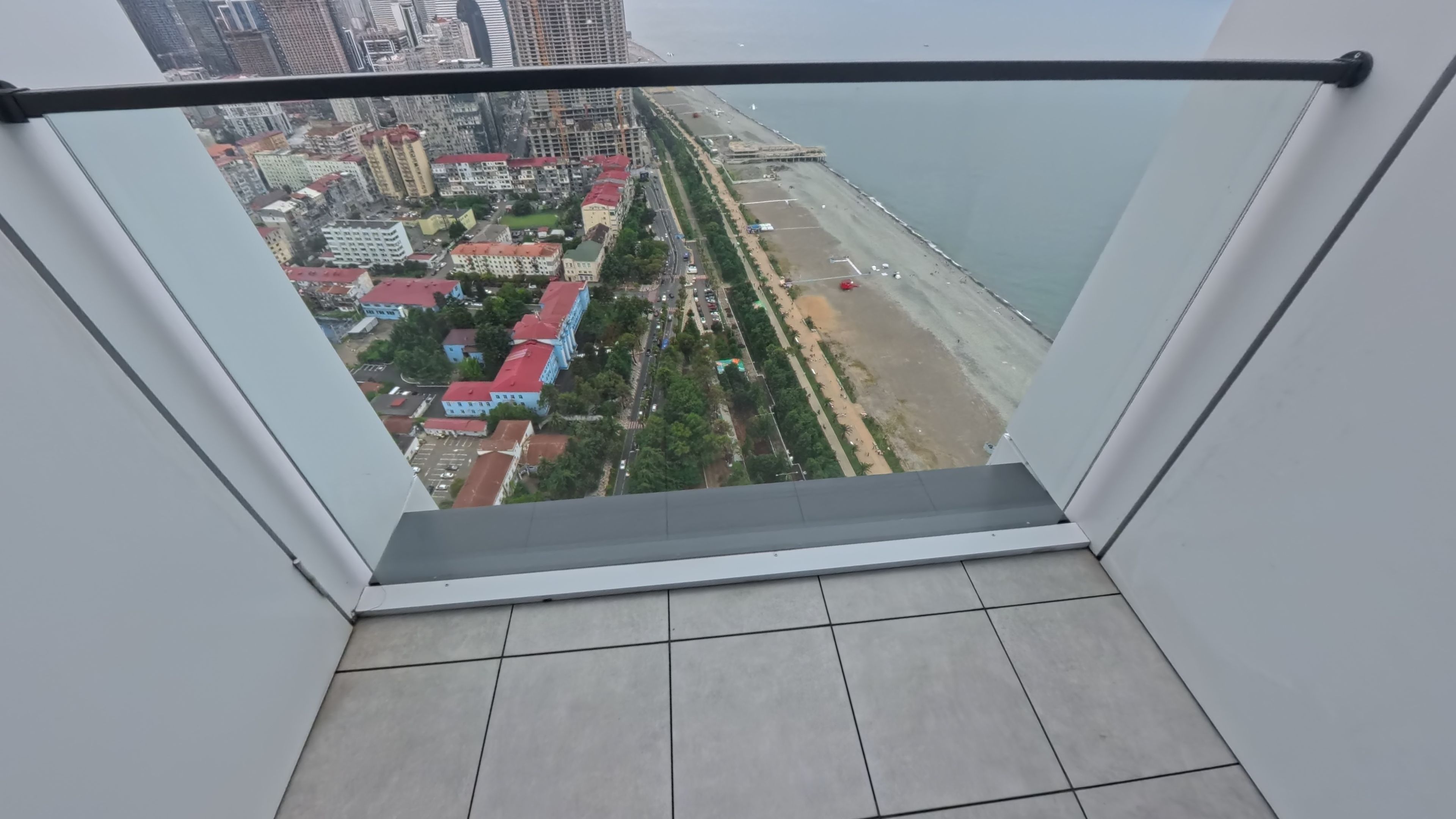 Vista desde un balcón con barandilla metálica, que da a una playa y un paisaje urbano con edificios y calles.