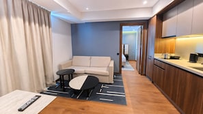 Habitación de hotel moderna con sofá, dos mesitas, un control remoto sobre una mesa y una zona de cocina con armarios y encimera.