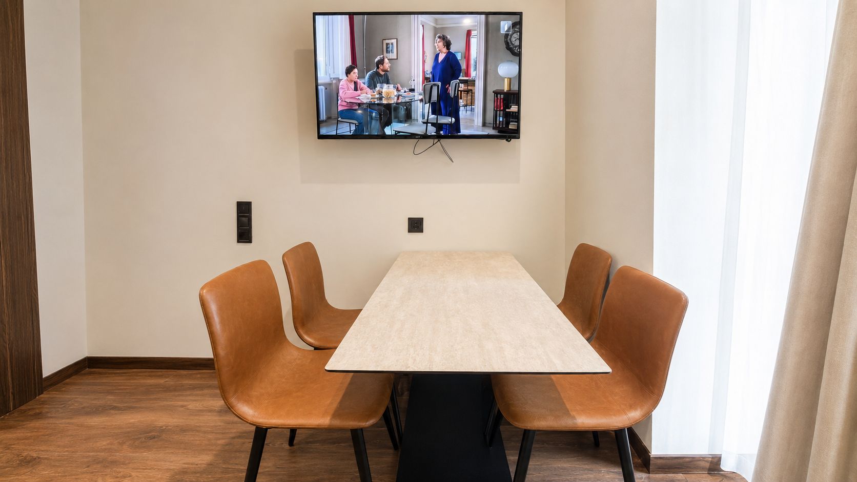 A conference room with a large table and four chairs, a flat-screen TV mounted on the wall, and a window with curtains.