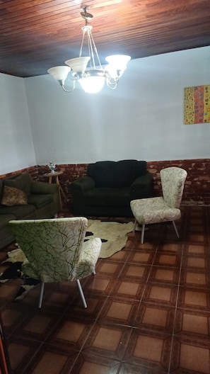 A living room with a green sofa, a patterned armchair, and a white armchair. A ceiling fan and a hanging light fixture are visible. The room has a tiled floor and a brick wall. - House in Saquarema, spacious with games room and gourmet area!!! (Saquarema)