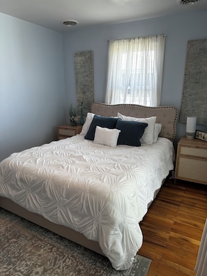 A bedroom with a large bed, wooden headboard, bedside tables, a window with sheer curtains, and a patterned rug. - Potomac River 2 Bedroom Cottage in Piney Point with Private Beach (Piney Point)