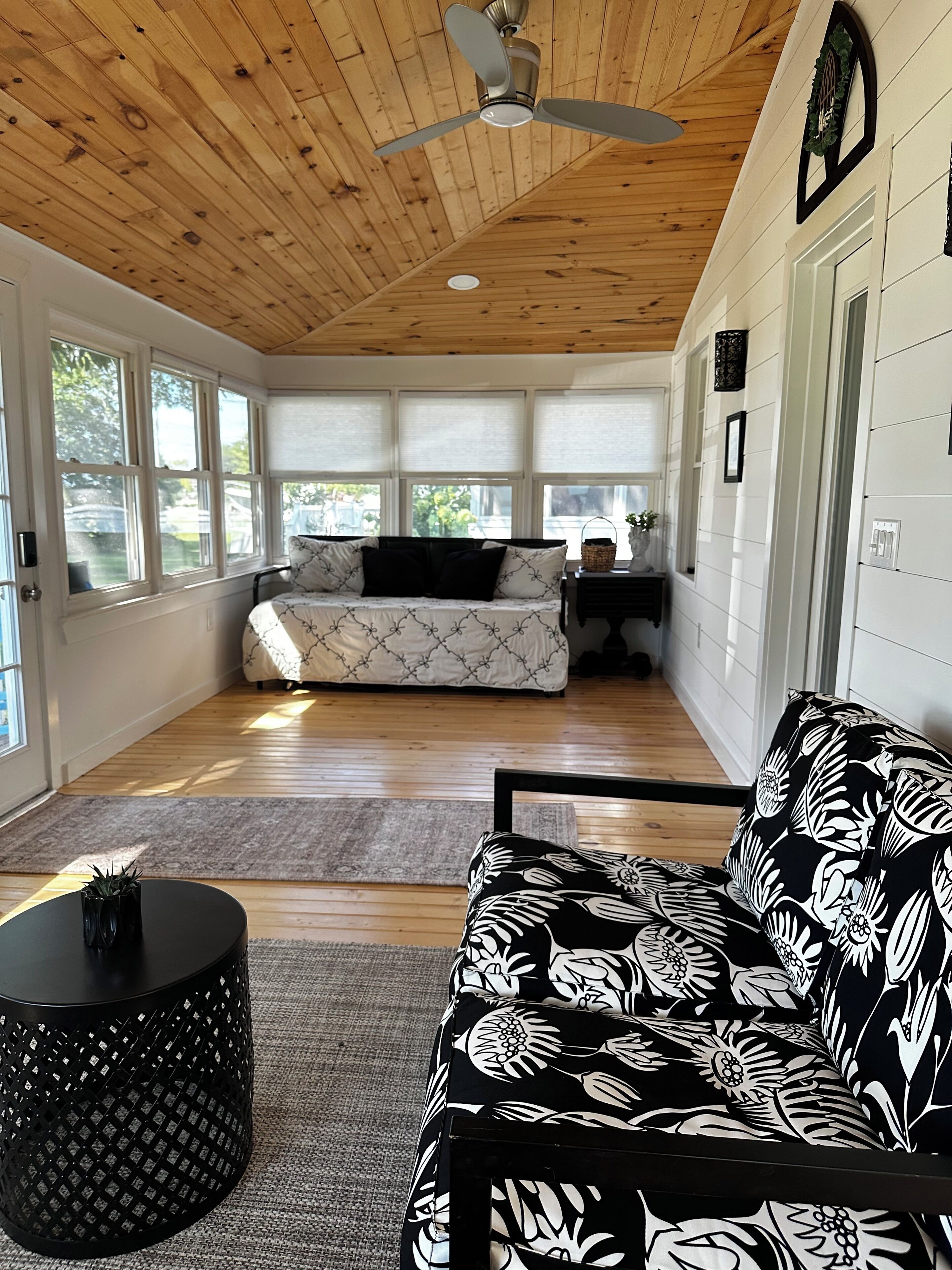 A room with a wooden ceiling, a ceiling fan, a sofa with a floral pattern, a round black table, and a window with a view of greenery.