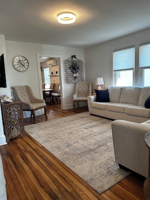 A living room with a wooden floor, a beige sofa, a round coffee table, and a wall clock. - Potomac River 2 Bedroom Cottage in Piney Point with Private Beach (Piney Point)