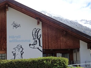 A building with a white exterior and wooden beams, featuring a mural with a butterfly, a dog, and the text 'Hörzi willkommen'. - Carmen's Oase, (Pany), by Interhome (Pany)