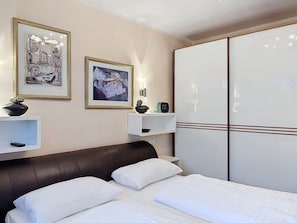 Ein Hotelzimmer mit einem Bett, einem Kleiderschrank, zwei gerahmten Kunstwerken, einer Pflanze auf einem Regal und einem Nachttisch.