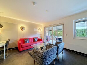 Ein modernes Wohnzimmer mit einem roten Sofa, gemusterten Sesseln, einem Glastisch und einem großen Fenster mit Jalousien.