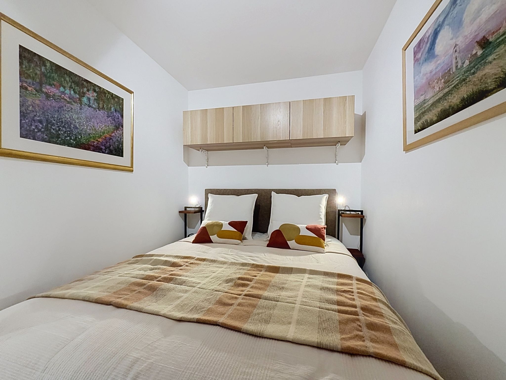 Un dormitorio con una cama, cabecera, dos mesitas de noche, un armario de madera sobre la cama y dos cuadros enmarcados en las paredes.
