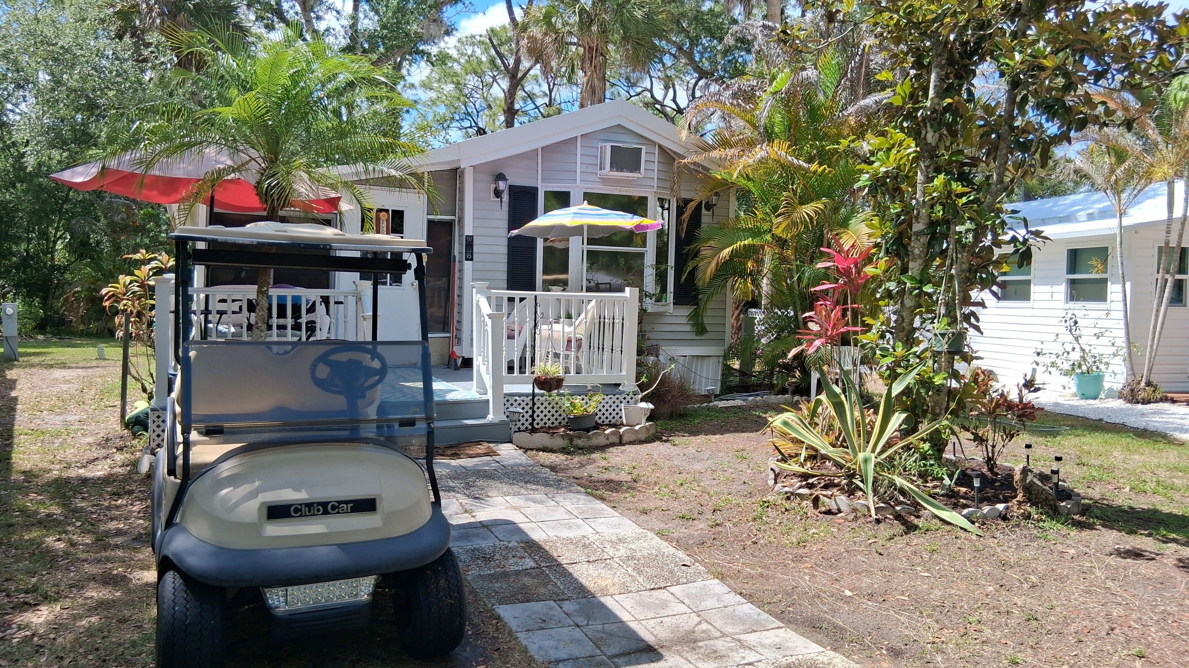 Un golf cart parcheggiato di fronte a una casa con veranda, circondata da piante tropicali e un cielo sereno.