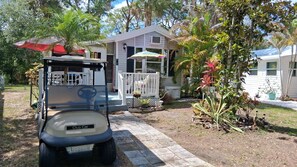 A golf cart parked in front of a house with a porch, surrounded by tropical plants and a clear sky. - Charming cottage getaway (Venice)