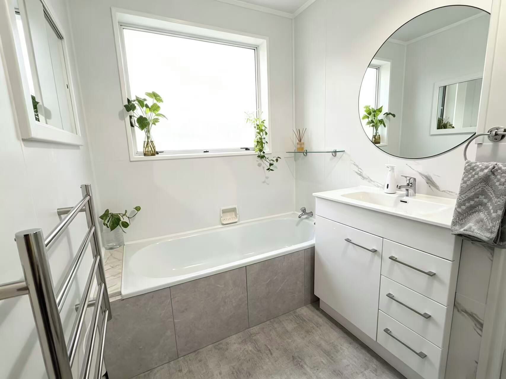 Een moderne badkamer met een badkuip, een glazen plank met planten, een grote spiegel en een witte wastafel met lades.