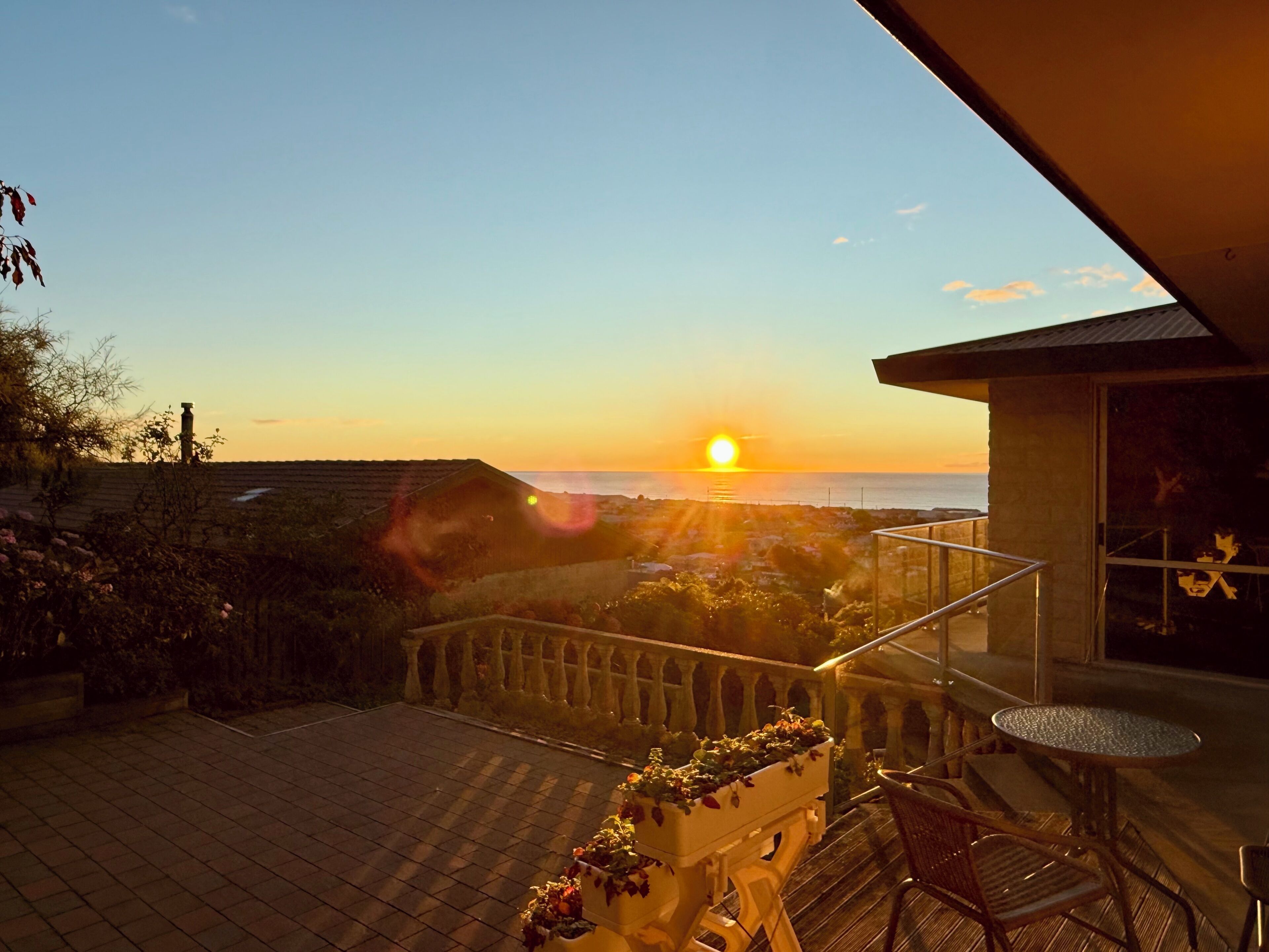 Ein Balkon mit Blick auf den Sonnenuntergang über dem Meer, einer Topfpflanze und einem Tisch mit Stühlen.