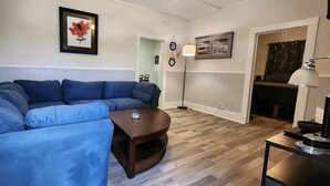 A living room with a blue sectional sofa, a small coffee table, and a lamp. There is a framed picture on the wall and a door leading to another room.
