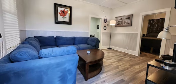 A living room with a blue sectional sofa, a small coffee table, and a lamp. There is a framed picture on the wall and a door leading to another room.