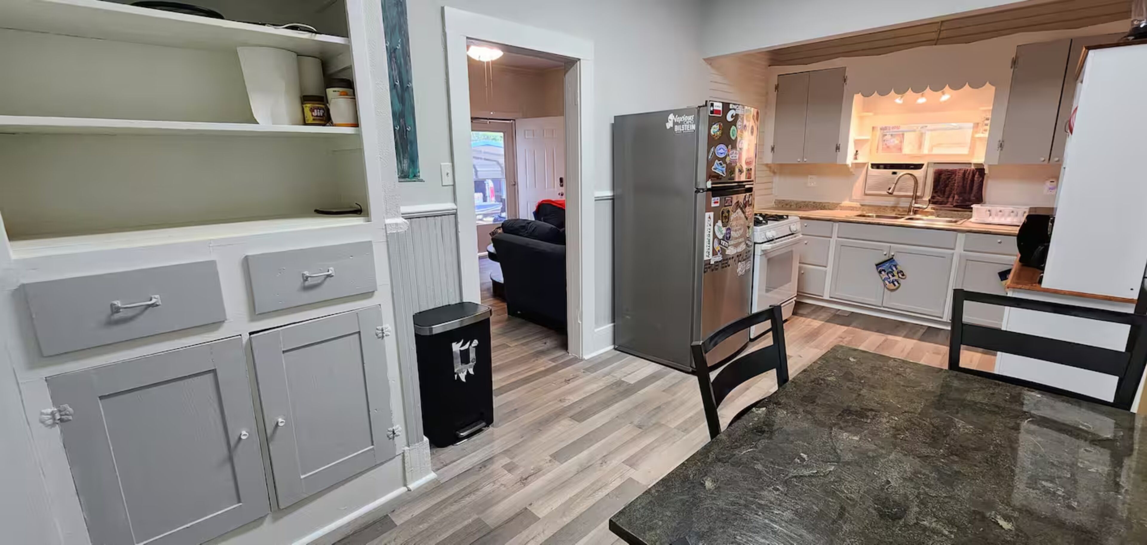 A compact kitchen with white cabinets, a black trash bin, a dining table, and a refrigerator.