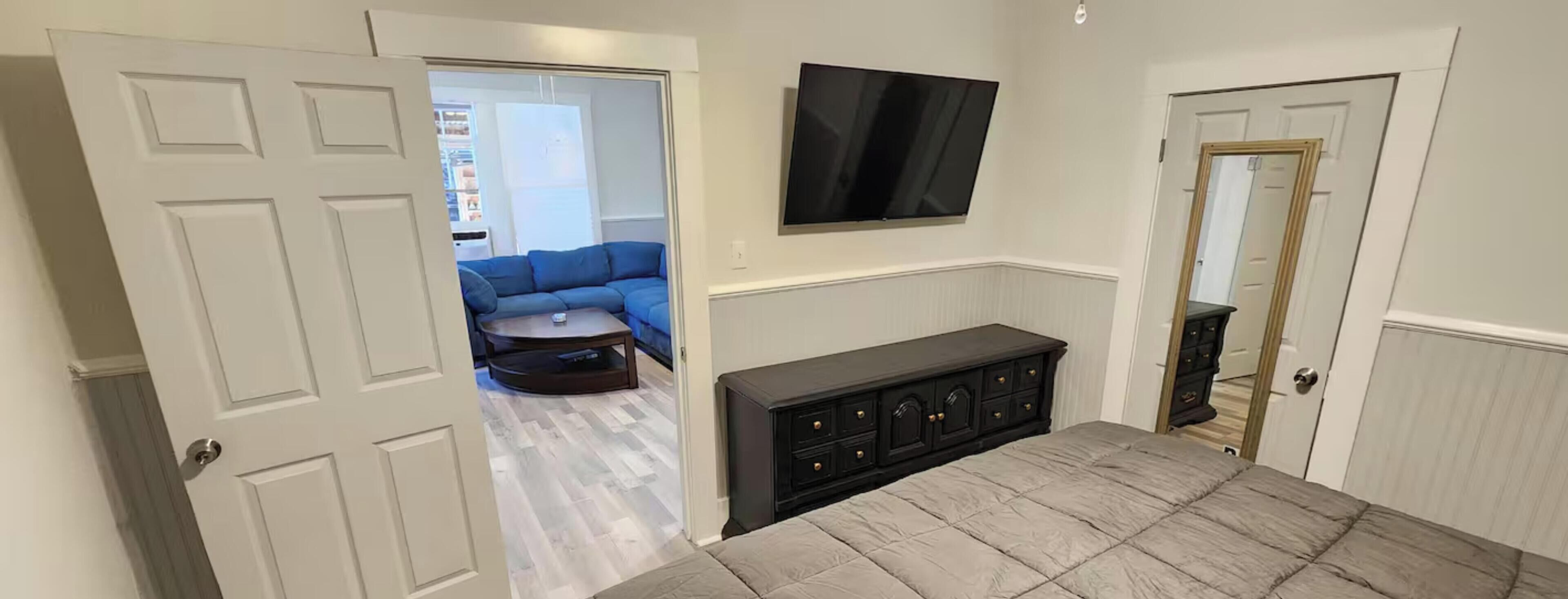 A room with a bed, a TV mounted on the wall, a dark-colored cabinet, and a door leading to another room.