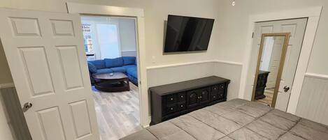 A room with a bed, a TV mounted on the wall, a dark-colored cabinet, and a door leading to another room.