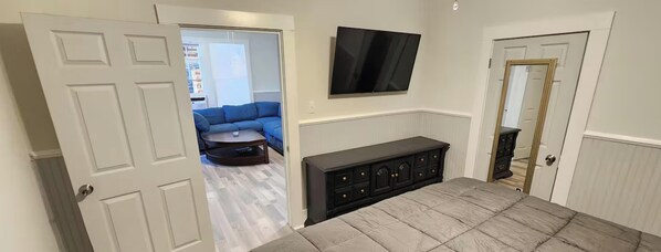A room with a bed, a TV mounted on the wall, a dark-colored cabinet, and a door leading to another room.