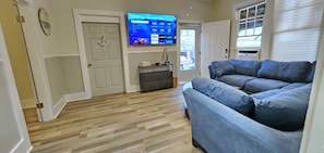 A living room with a blue sofa, a flat-screen TV mounted on the wall, a wooden floor, and a door with a white frame.