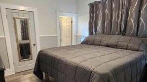 A bedroom with a bed, a bedside table, a nightstand, a mirror, and a door.